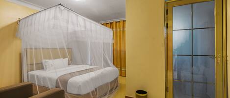 A hotel room with a bed under a mosquito net, a small trash bin, and a sliding glass door.