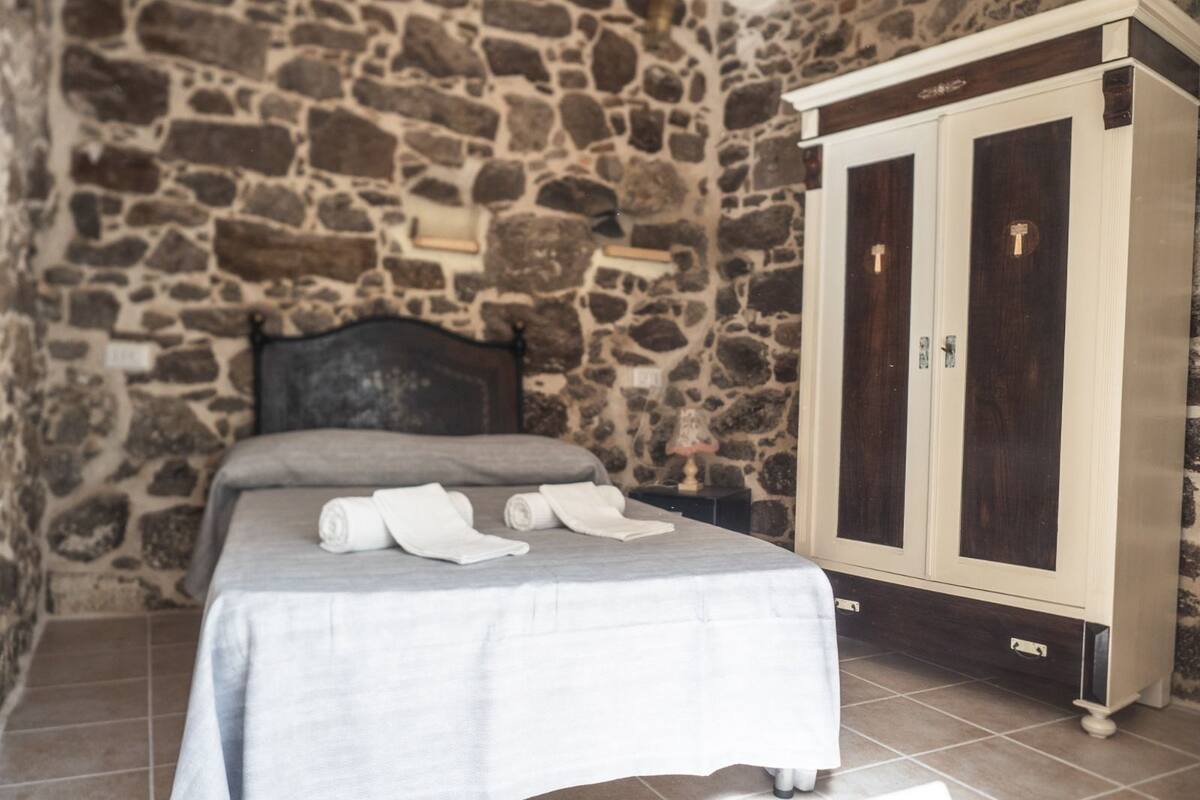 A bedroom with a stone wall, a bed with a grey bedspread, and a wooden wardrobe with two doors.