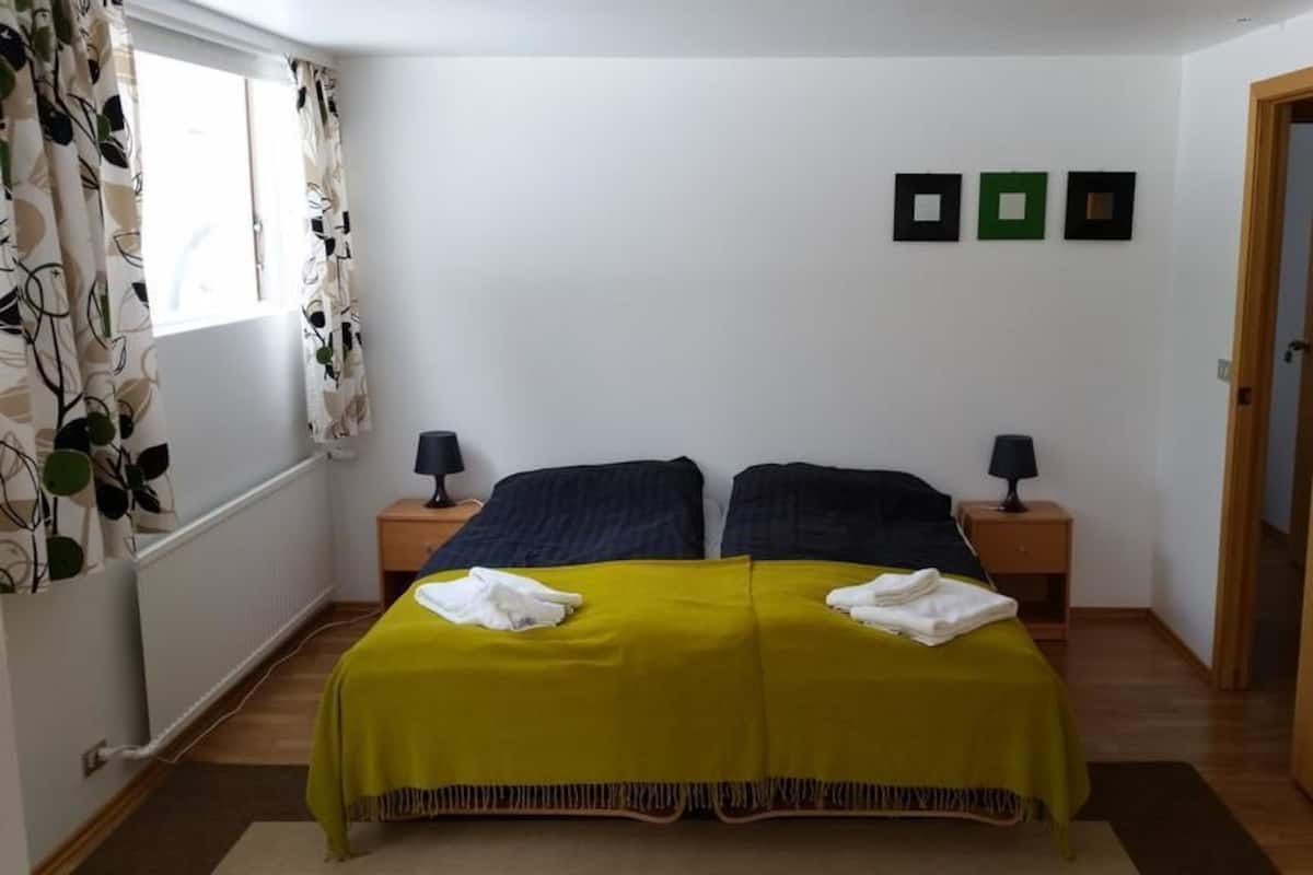 A bedroom with a double bed, two bedside tables with lamps, a wooden door, and a window with curtains.