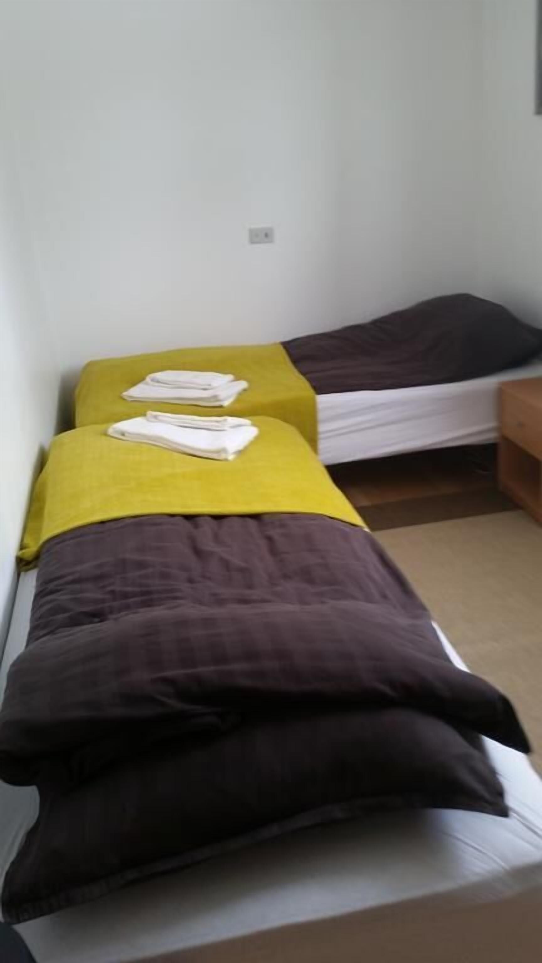 Dos camas individuales con ropa de cama amarilla y marrón, una mesita de noche de madera y una pared blanca con toma de corriente.