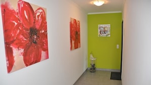 A hallway with a green wall, two large red floral paintings, a small white pedestal with a vase, and a door at the end.