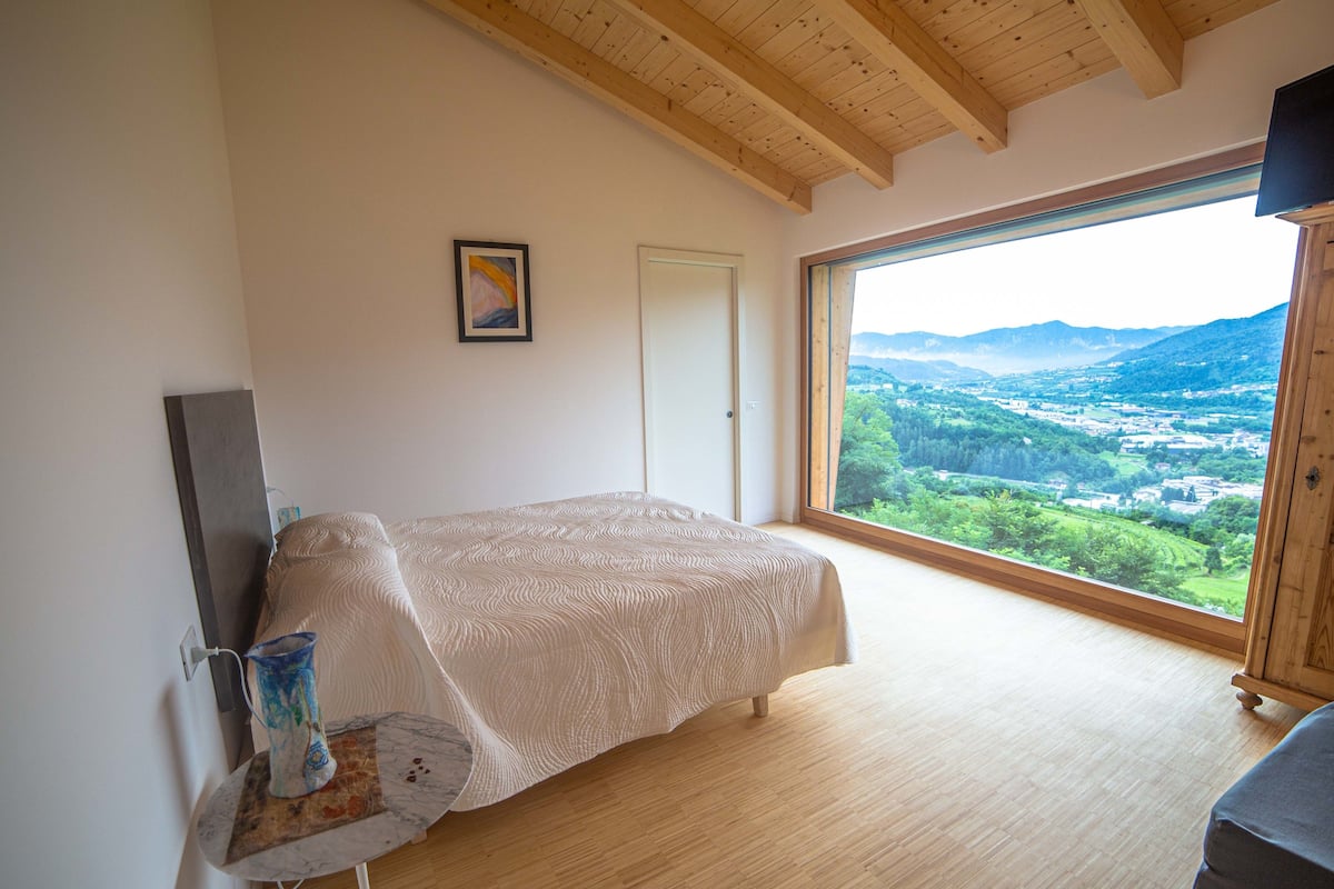 Une chambre à coucher avec un lit, un plancher en bois, une grande fenêtre donnant sur une vallée et un tableau accroché au mur.