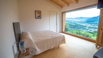 A bedroom with a bed, a wooden floor, a large window with a view of a valley, and a painting on the wall.