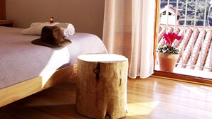 A bedroom with a wooden bed frame, a small wooden stool, a hat, and a potted plant on a balcony with a view of a forested area. - Hotel La Tinensa (La Pobla de Benifassa)