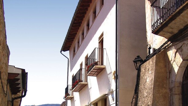 A narrow street lined with multi-story buildings, featuring balconies with flower boxes and potted plants. - Hotel La Tinensa (La Pobla de Benifassa)