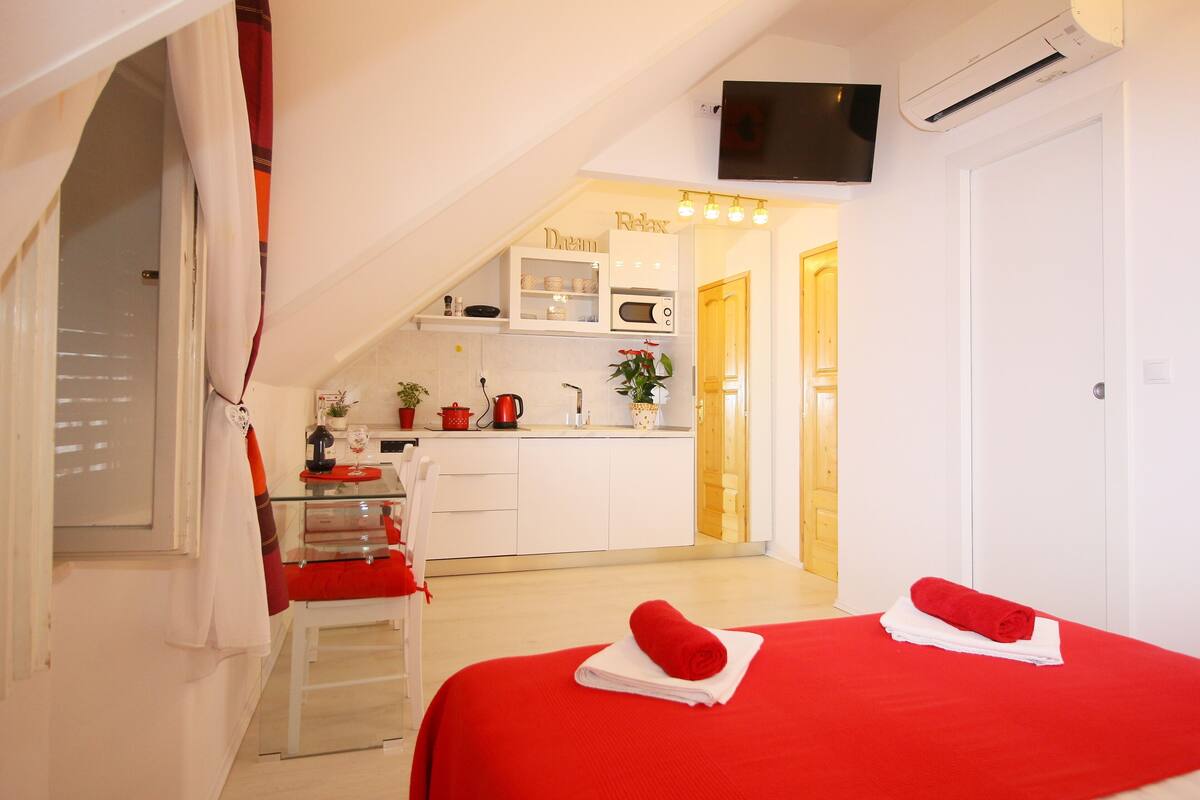 A compact bedroom with a red bedspread, a small kitchenette, and a TV mounted on the wall.