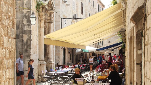 A narrow street with outdoor seating and tables, stone buildings, and a clock. - Melek Accommodations (Dubrovnik)