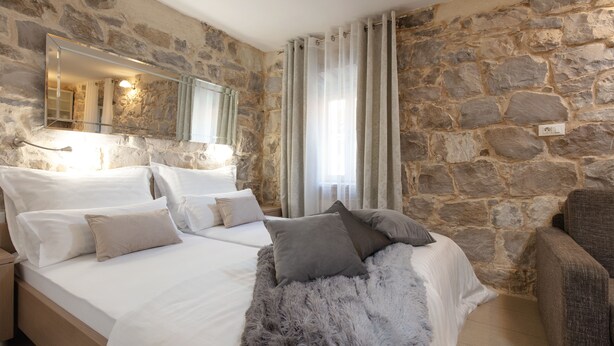Un dormitorio con pared de piedra, una cama con ropa de cama blanca, un plaid gris suave y una silla.
