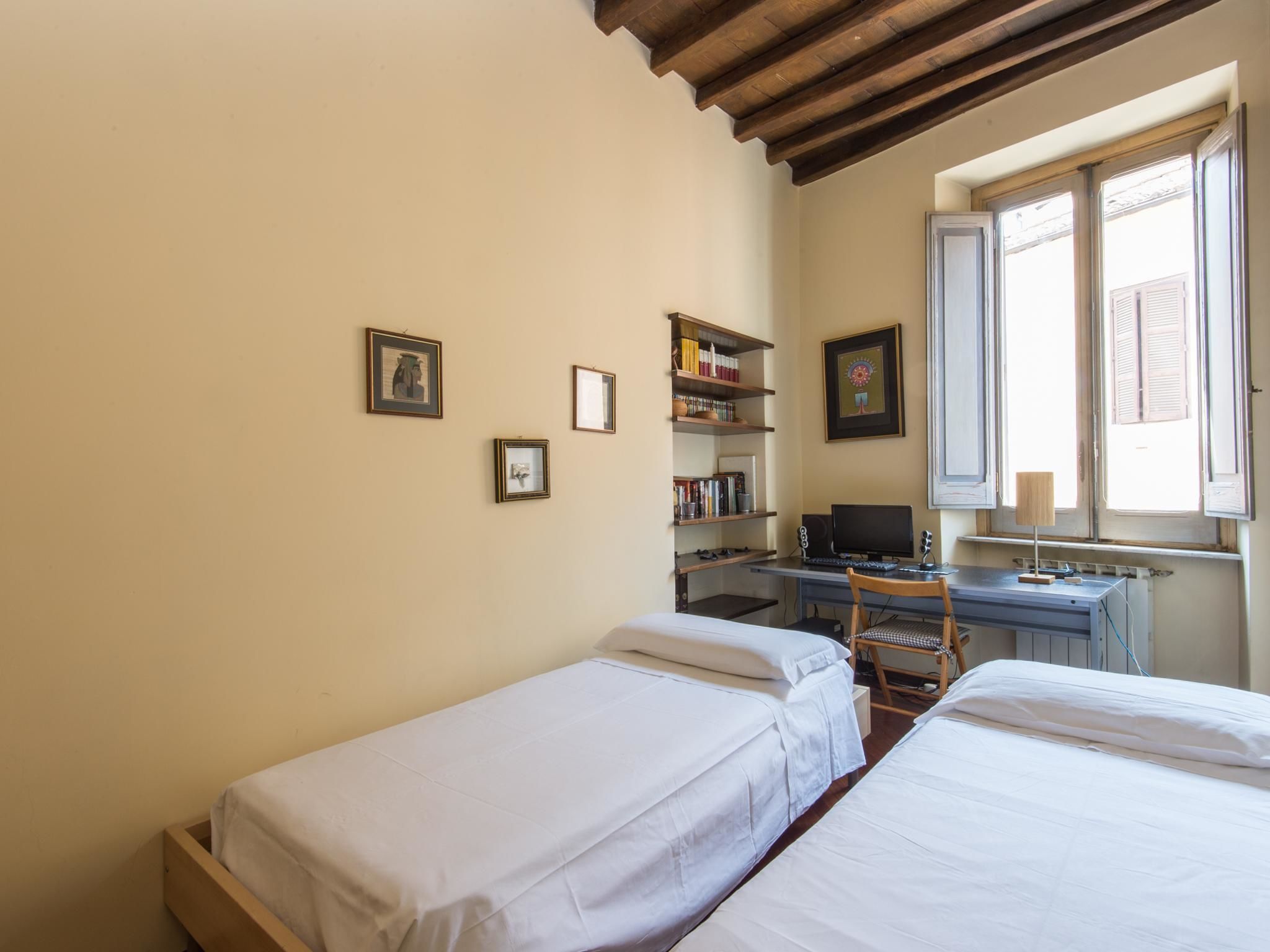 Un dormitorio con dos camas individuales, un escritorio con computadora, una estantería y una ventana con contraventanas.