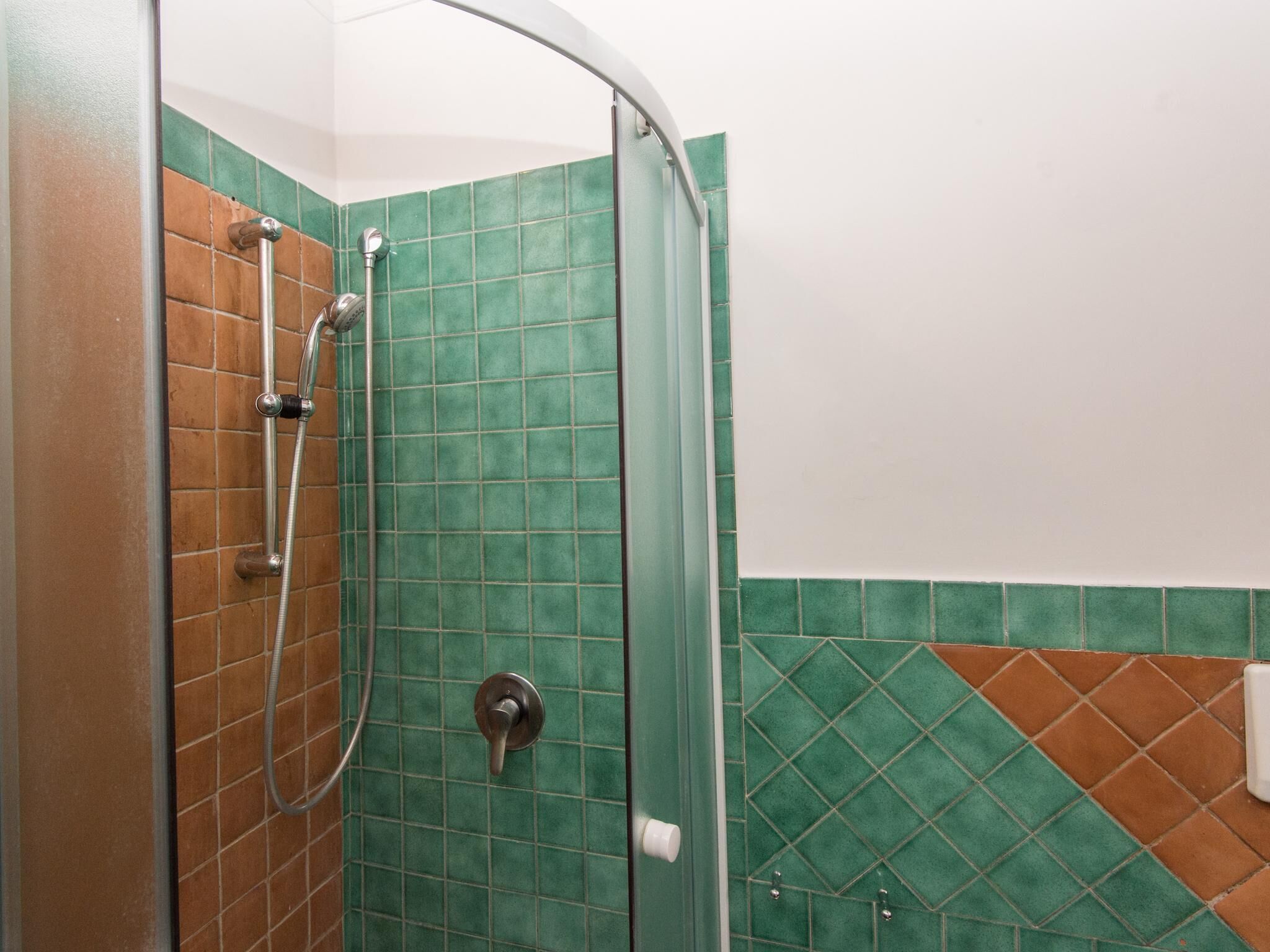 A shower area with a curved glass door, green and brown tiles, and a handheld showerhead.