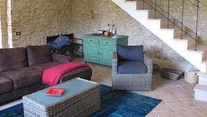 A living room with a stone wall, a staircase, a green cabinet, a brown sofa, a blue armchair, and a wicker coffee table.