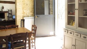A dining area with a stone wall, a wooden table, and chairs. There is a glass door leading to a kitchen with a white refrigerator and a wire mesh cabinet.