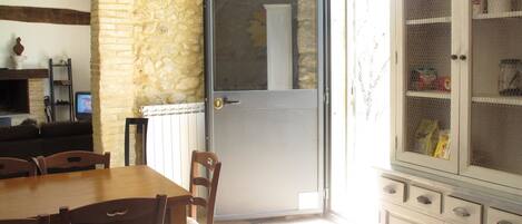 A dining area with a stone wall, a wooden table, and chairs. There is a glass door leading to a kitchen with a white refrigerator and a wire mesh cabinet.