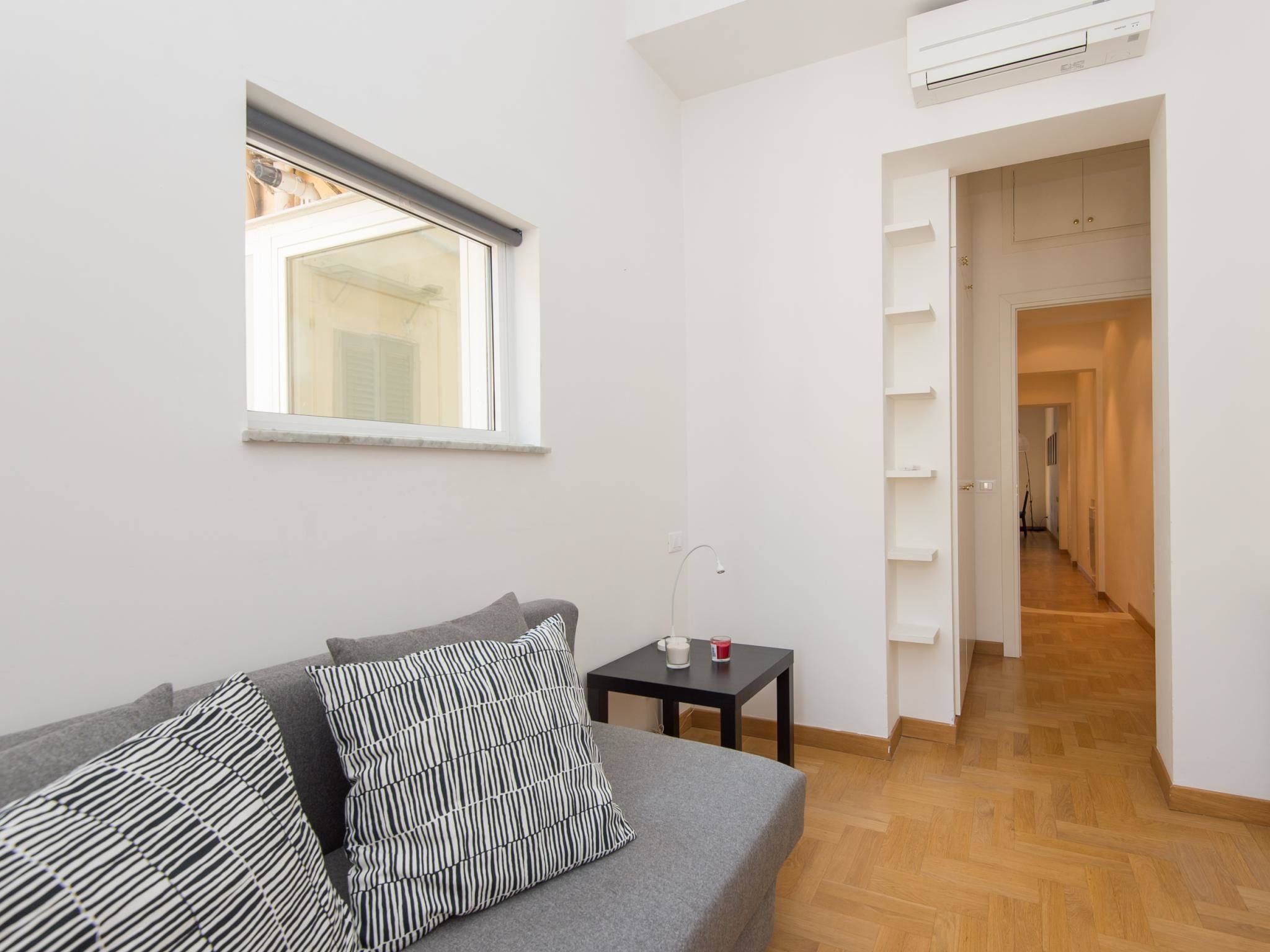 Ein modernes, minimalistisches Zimmer mit einem grauen Sofa, gestreiften Kissen, einem kleinen Tisch mit einer Tasse und einem Fenster mit Blick auf ein Gebäude.