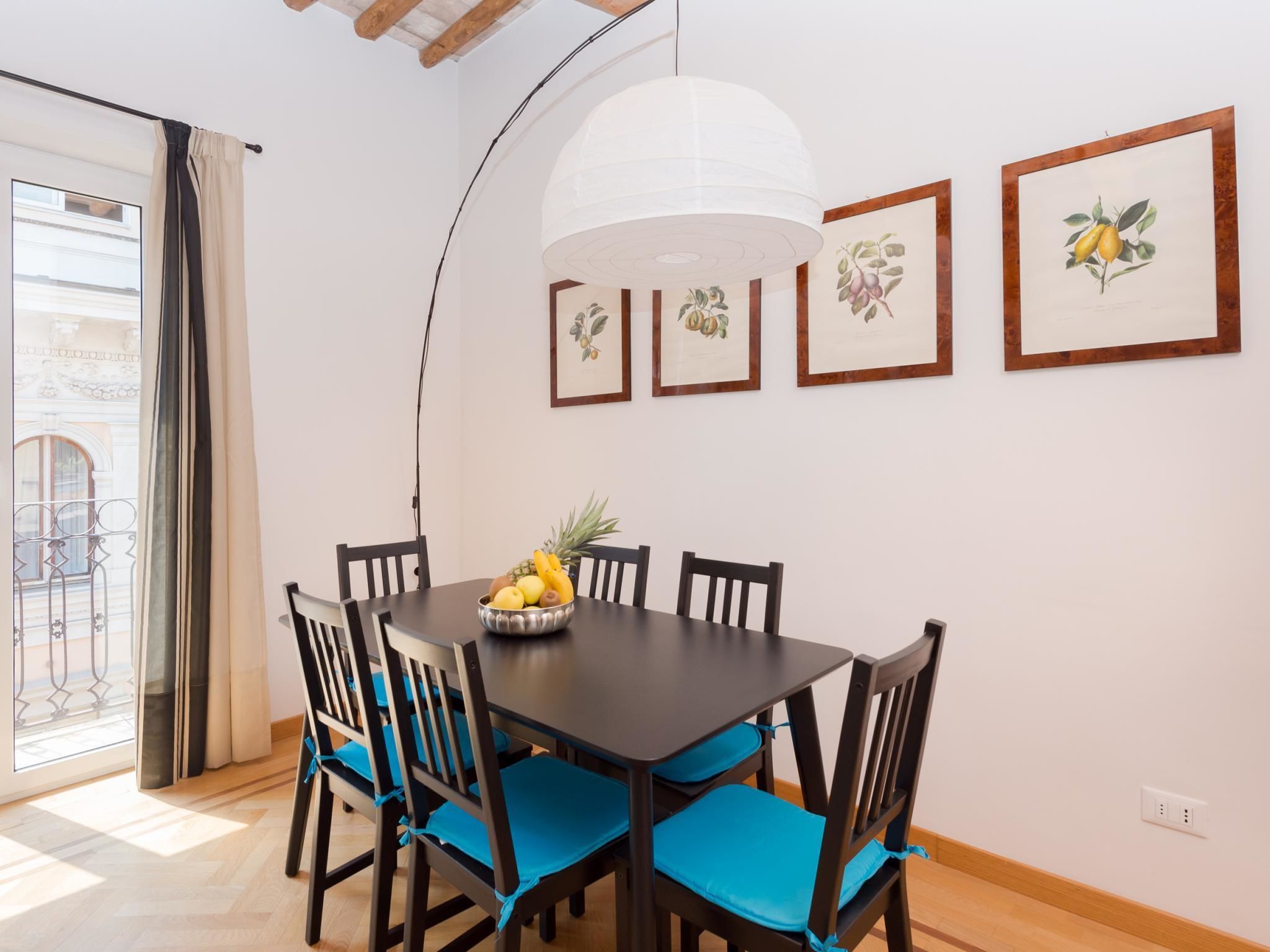 A dining area with a dark wooden table, blue cushioned chairs, and a fruit bowl. There are framed botanical prints on the wall and a white pendant light above the table.
