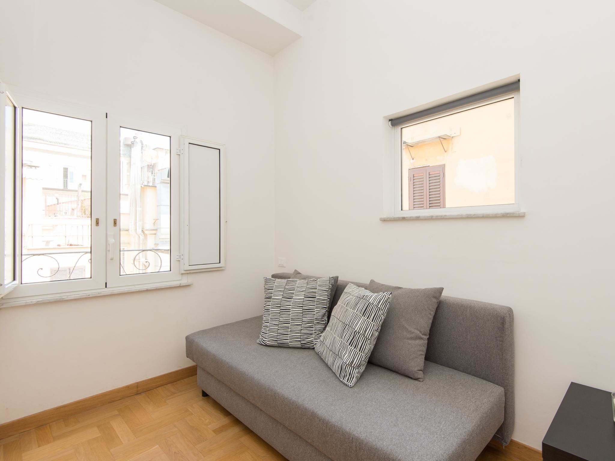 Una habitación con un sofá gris, dos almohadas estampadas, una mesita y dos ventanas con contraventanas.