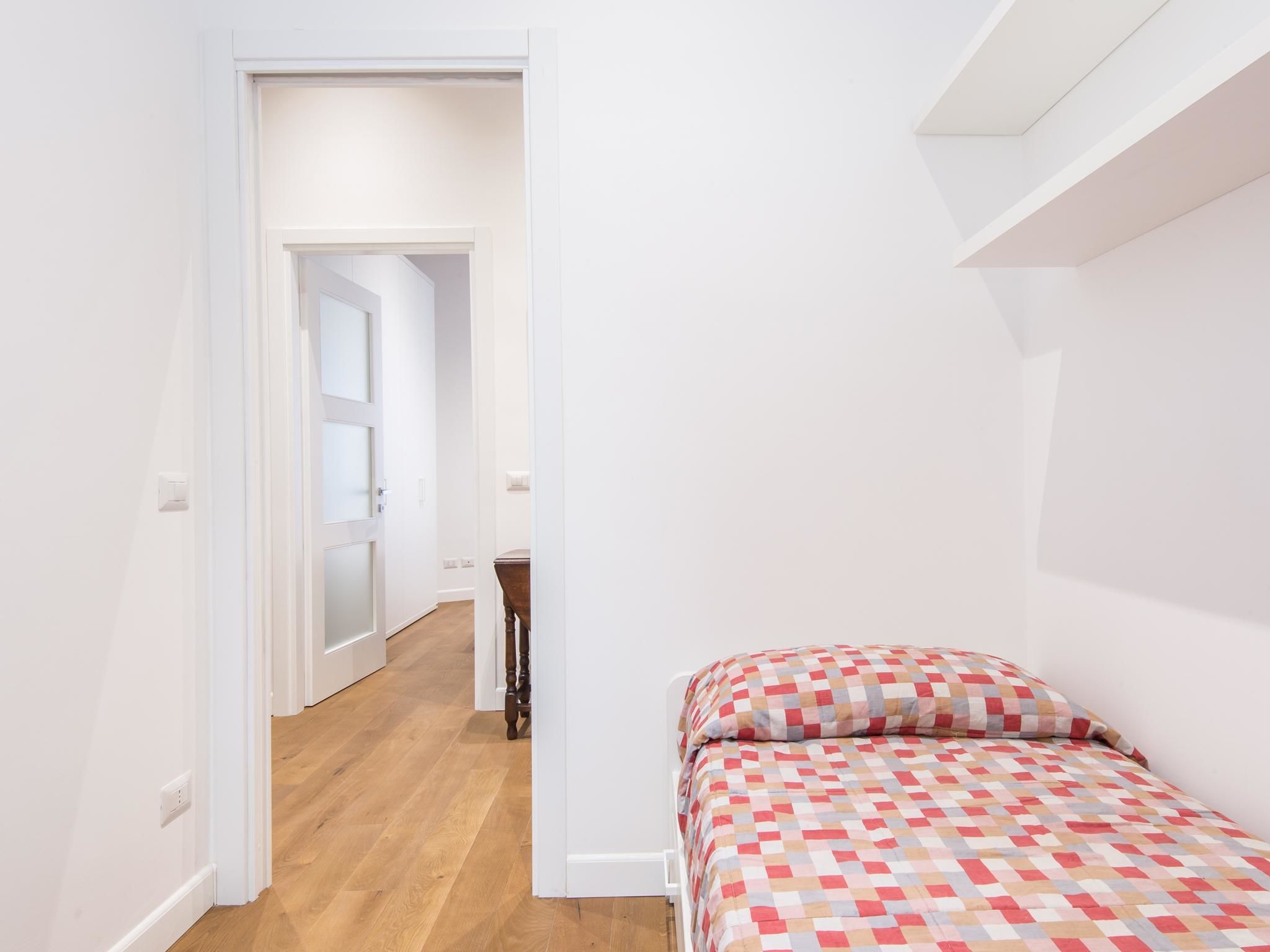 A small, brightly lit room with a wooden floor, a single bed with a checkered bedspread, and a white door leading to another room.