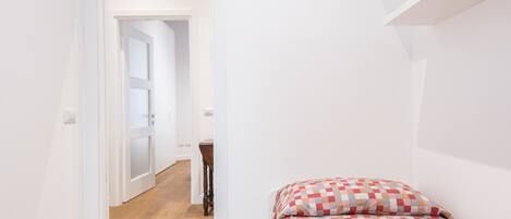 A small, brightly lit room with a wooden floor, a single bed with a checkered bedspread, and a white door leading to another room.