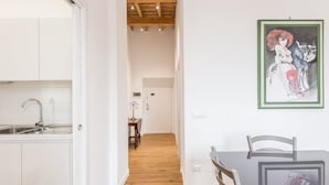 A modern kitchen with white cabinets and a wooden floor. There is a dining area with a table and chairs. A painting is visible on the wall.