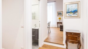 A hallway leading to a bathroom with a sink and mirror, a wooden floor, and a small table with a lamp.