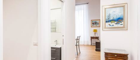 A hallway leading to a bathroom with a sink and mirror, a wooden floor, and a small table with a lamp.