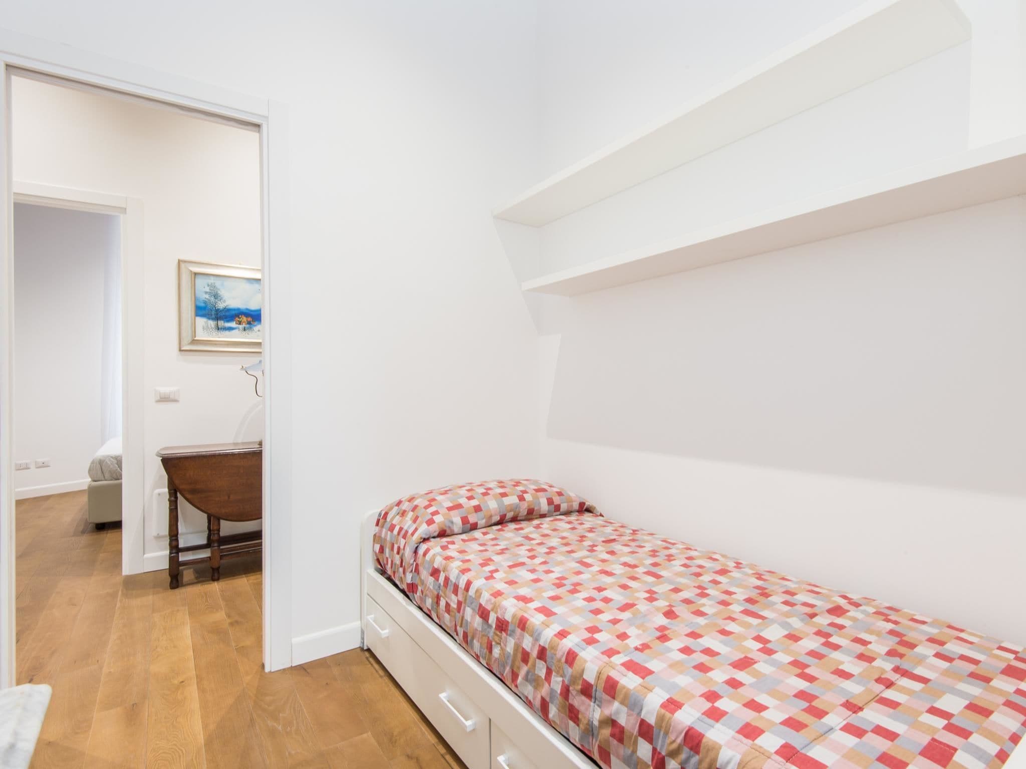 Une petite chambre avec un lit simple, une couverture à carreaux, un bureau en bois et un tableau accroché au mur.