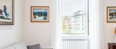 A living room with a white sofa, a radiator, and a window with a view of buildings.