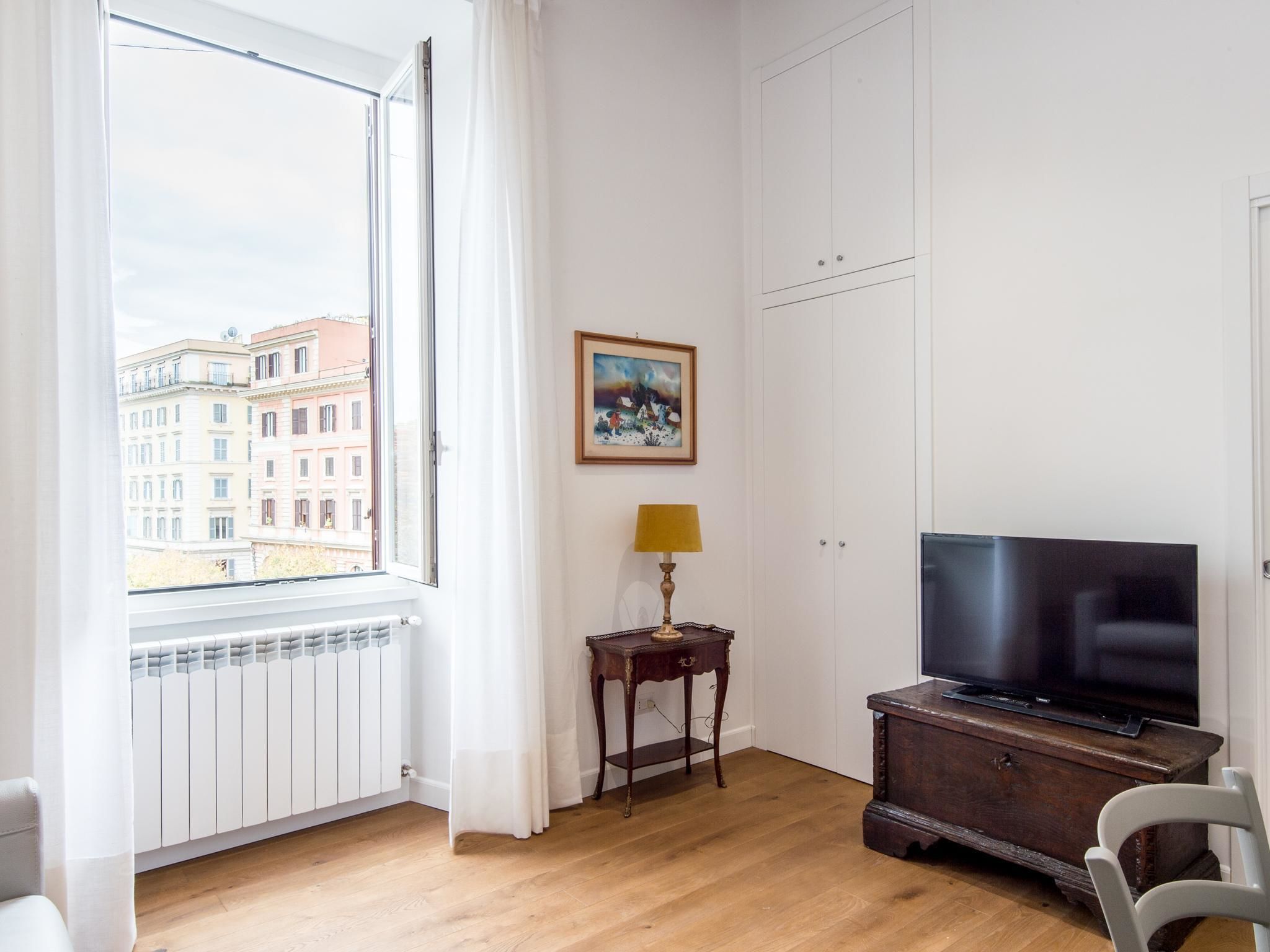 A room with a large window, a radiator, a wooden cabinet with a television, a small table with a lamp, and a painting on the wall.