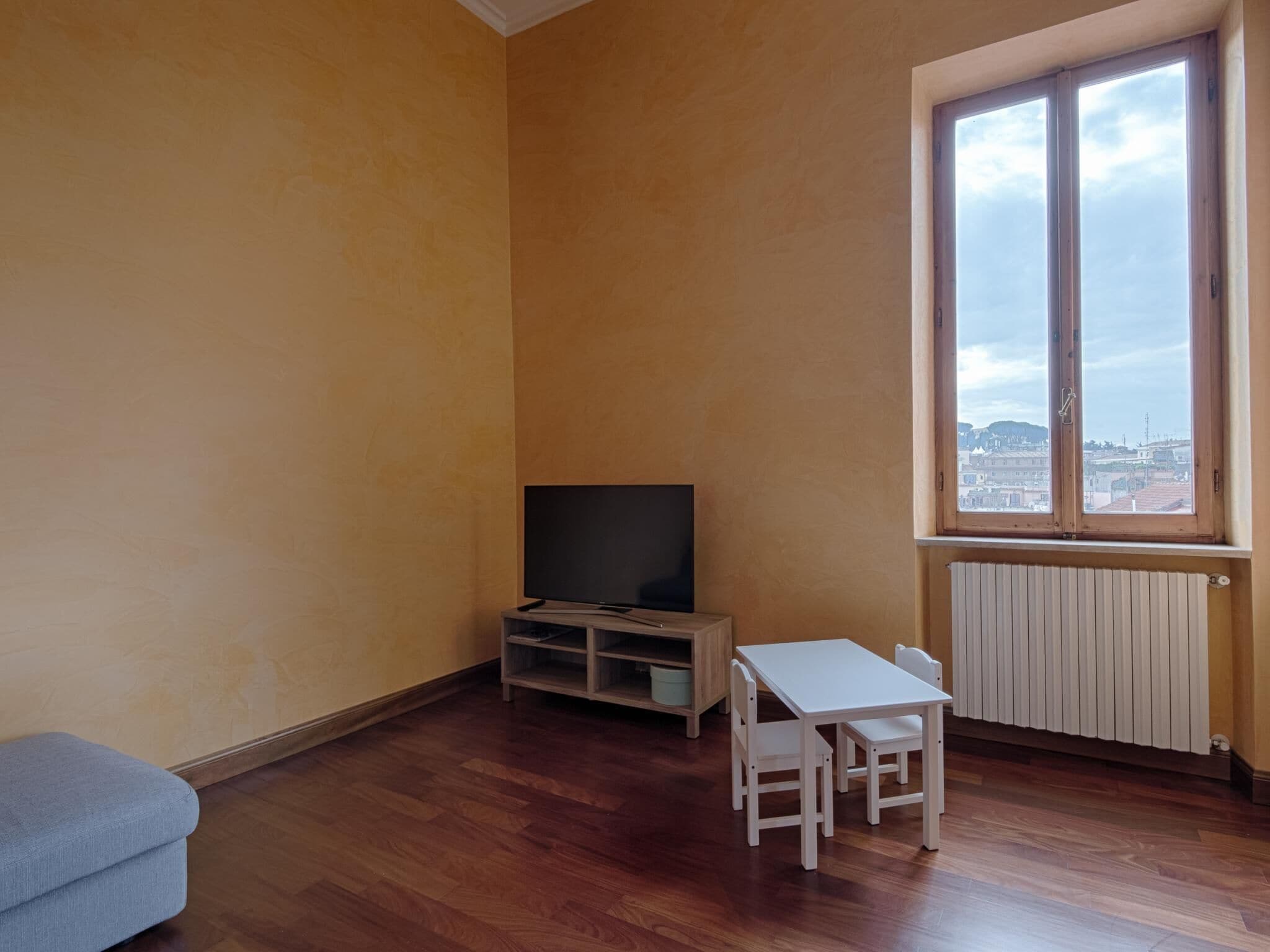 Une pièce avec un plancher en bois, un téléviseur posé sur un meuble, une table blanche avec des chaises et une fenêtre donnant sur des immeubles.