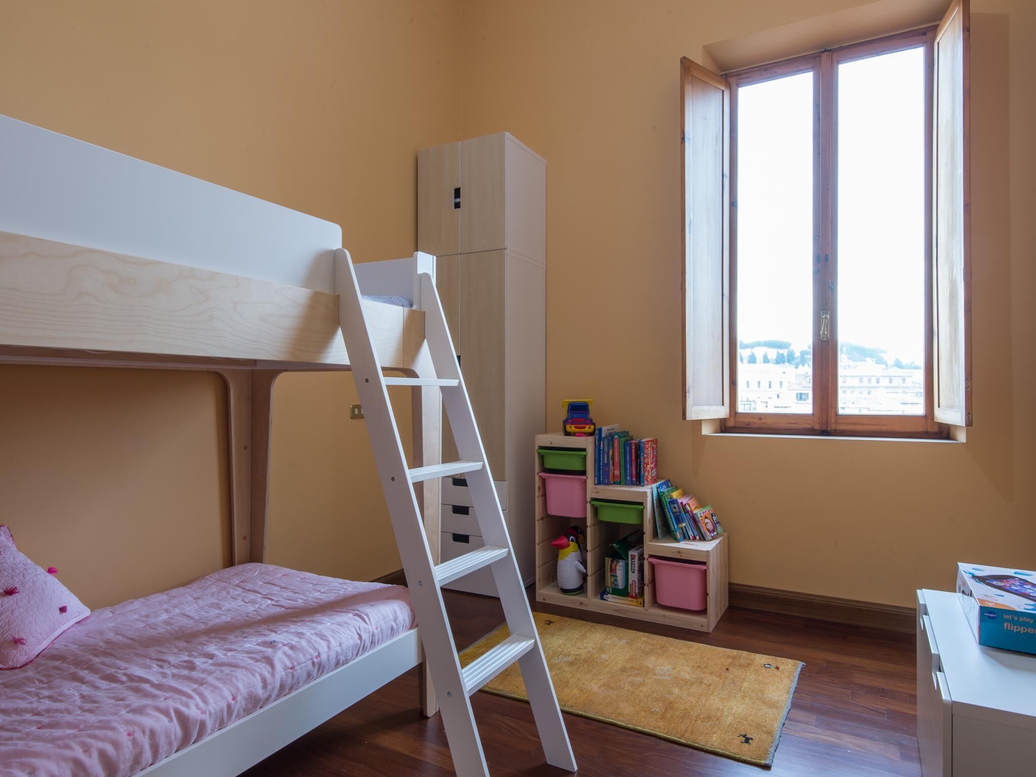 A child's bedroom with a bunk bed, a ladder, a wardrobe, a window with a view, and a toy storage unit.