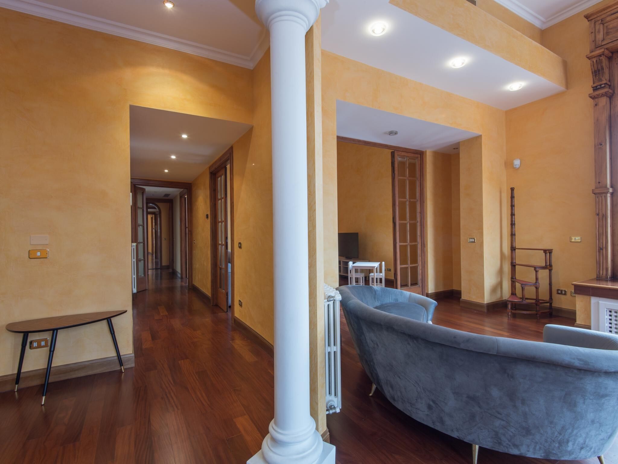 A spacious hallway with wooden flooring, a large white column, and a grey upholstered sofa.