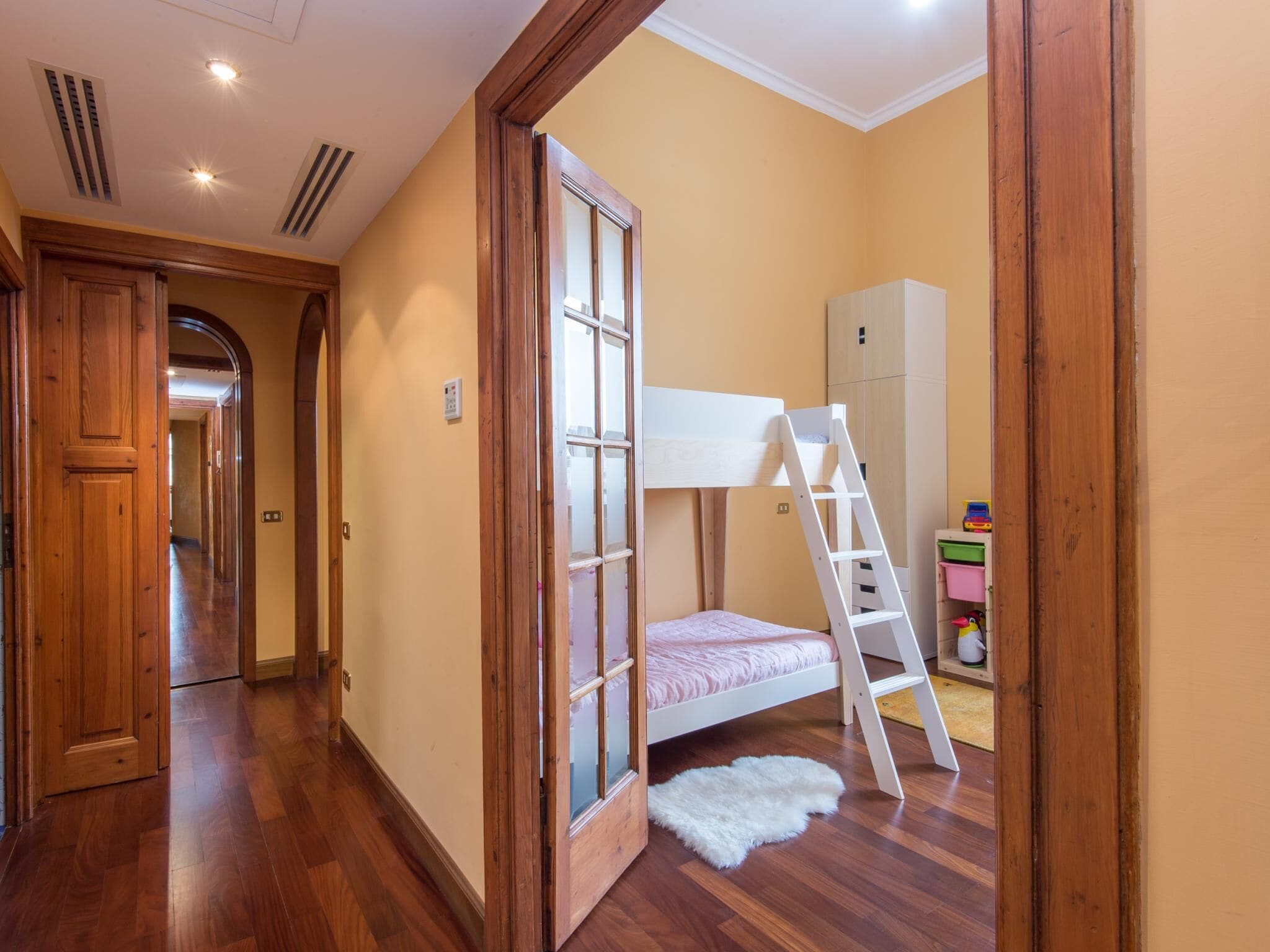 Una habitación con literas, suelo de madera, una escalera blanca, un armario y una puerta que da a otra habitación.