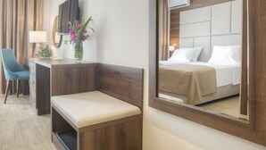Habitación de hotel con un espejo grande, una cama con ropa blanca, un escritorio con una silla, un pequeño banco y un florero con flores.