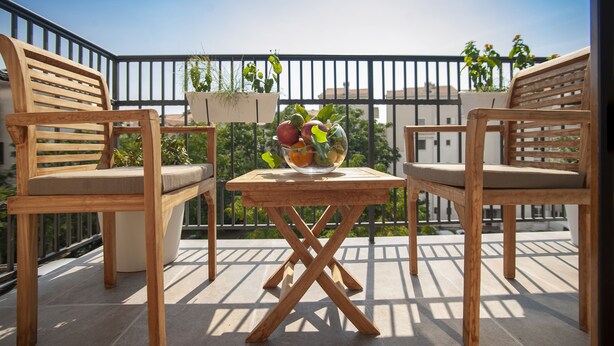 Un balcón de madera con dos sillas, una mesa, una planta en maceta y un cuenco de fruta.