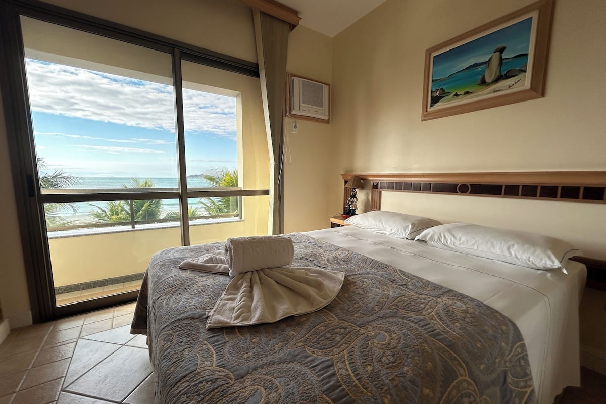A hotel room with a large bed, a painting on the wall, and a view of the ocean through a sliding door.