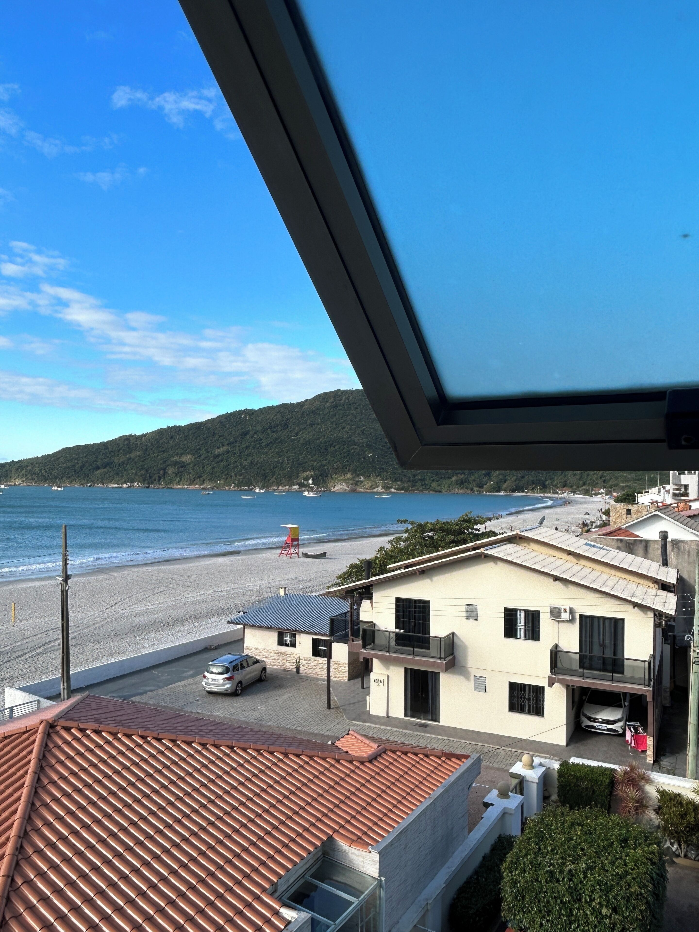 A coastal view from a balcony with a tiled roof, a beach, and a building with a car parked in front.