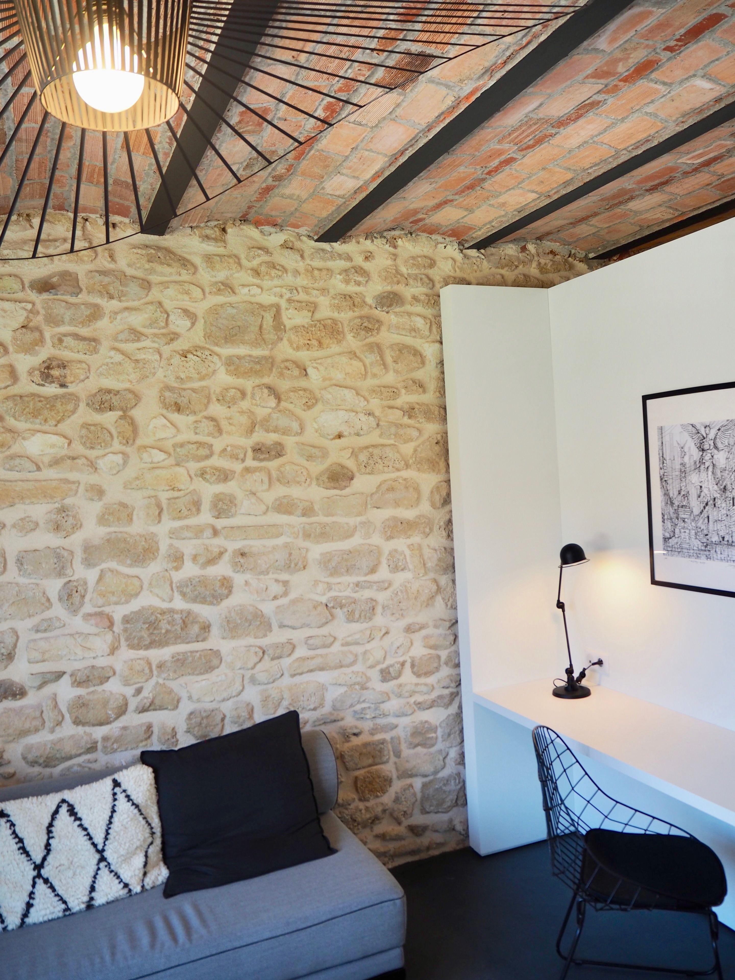 Une pièce avec un mur en pierre, un canapé gris avec un coussin noir, un lampadaire noir et une chaise noire avec un dossier en fil de fer.