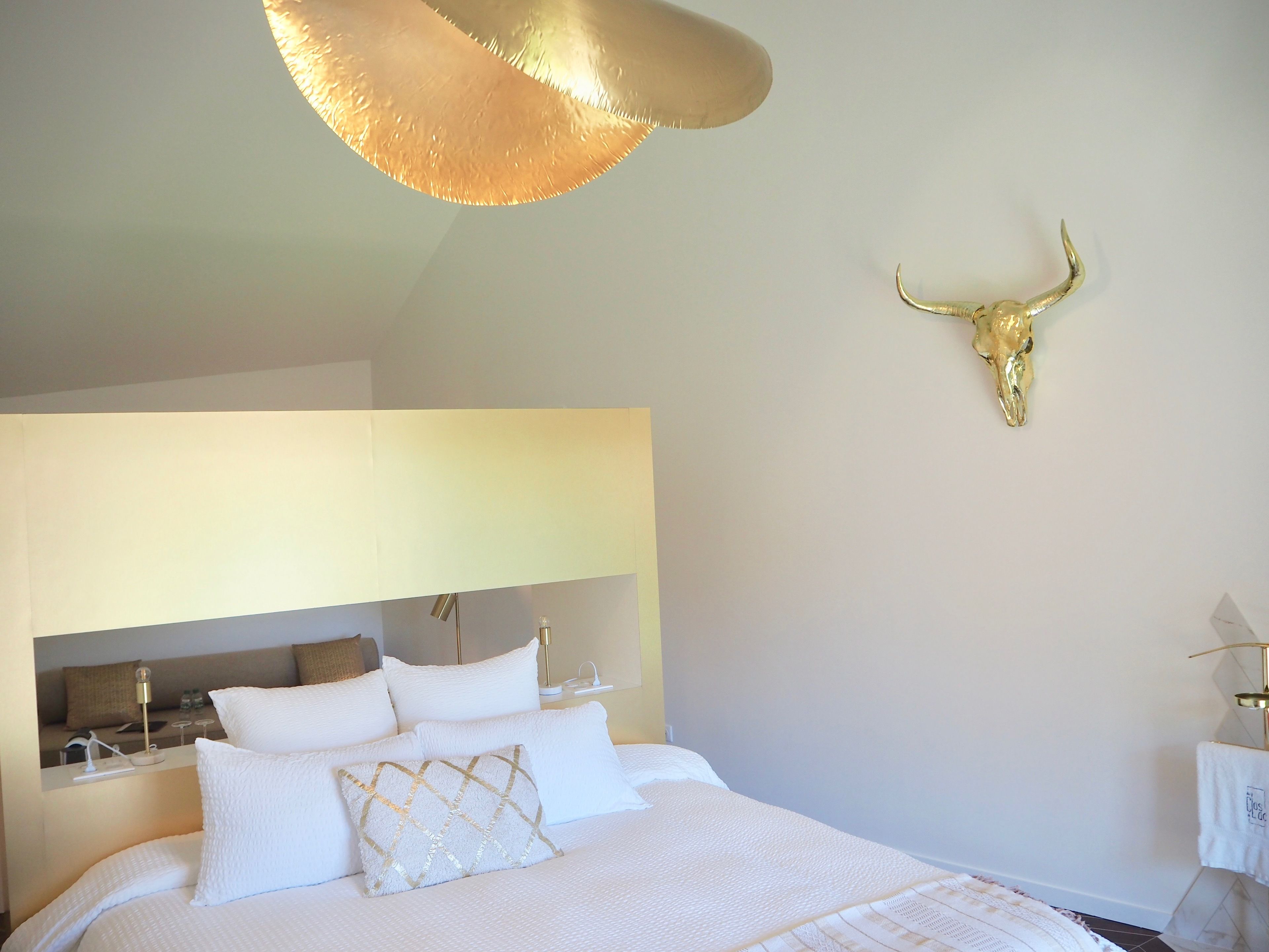 A modern hotel room with a large bed, white bedding, a gold-toned wall-mounted bull skull, and a unique ceiling light fixture.