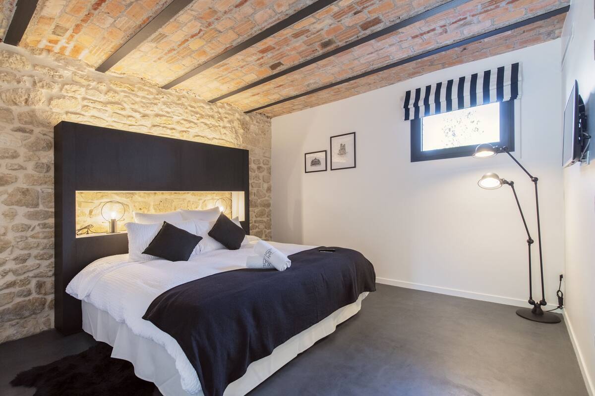 Quarto com parede de pedra, cama com roupa de cama branca, cabeceira preta, janela com valance listrado e luminária de mesa.