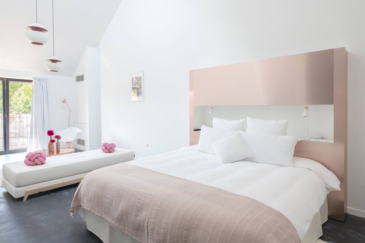 Um quarto moderno com uma cama grande, uma mesa de cabeceira, um banco com objetos decorativos e uma janela com cortinas.
