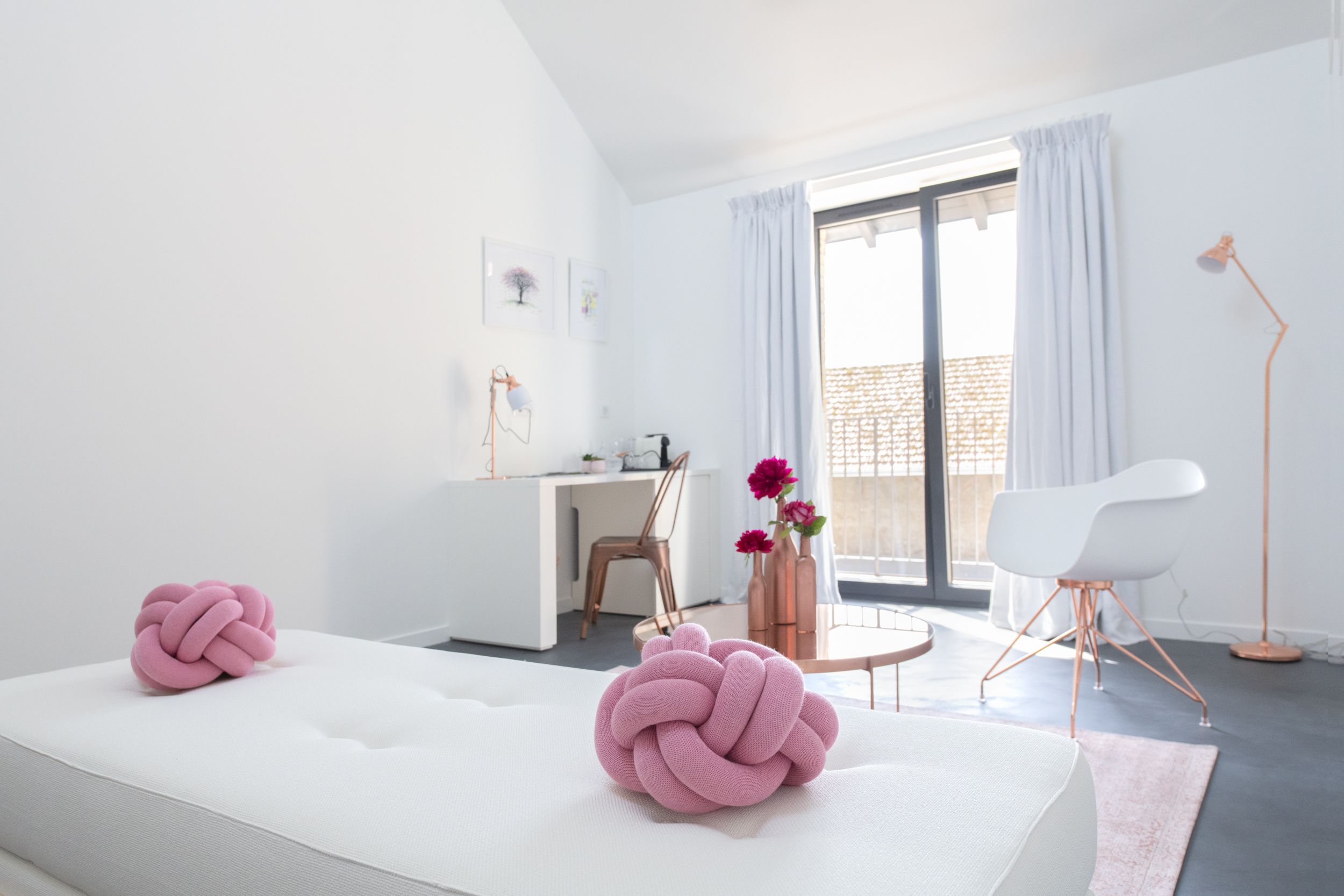 Un dormitorio moderno con una cama blanca, un escritorio y una silla. Hay almohadas decorativas rosas en la cama.