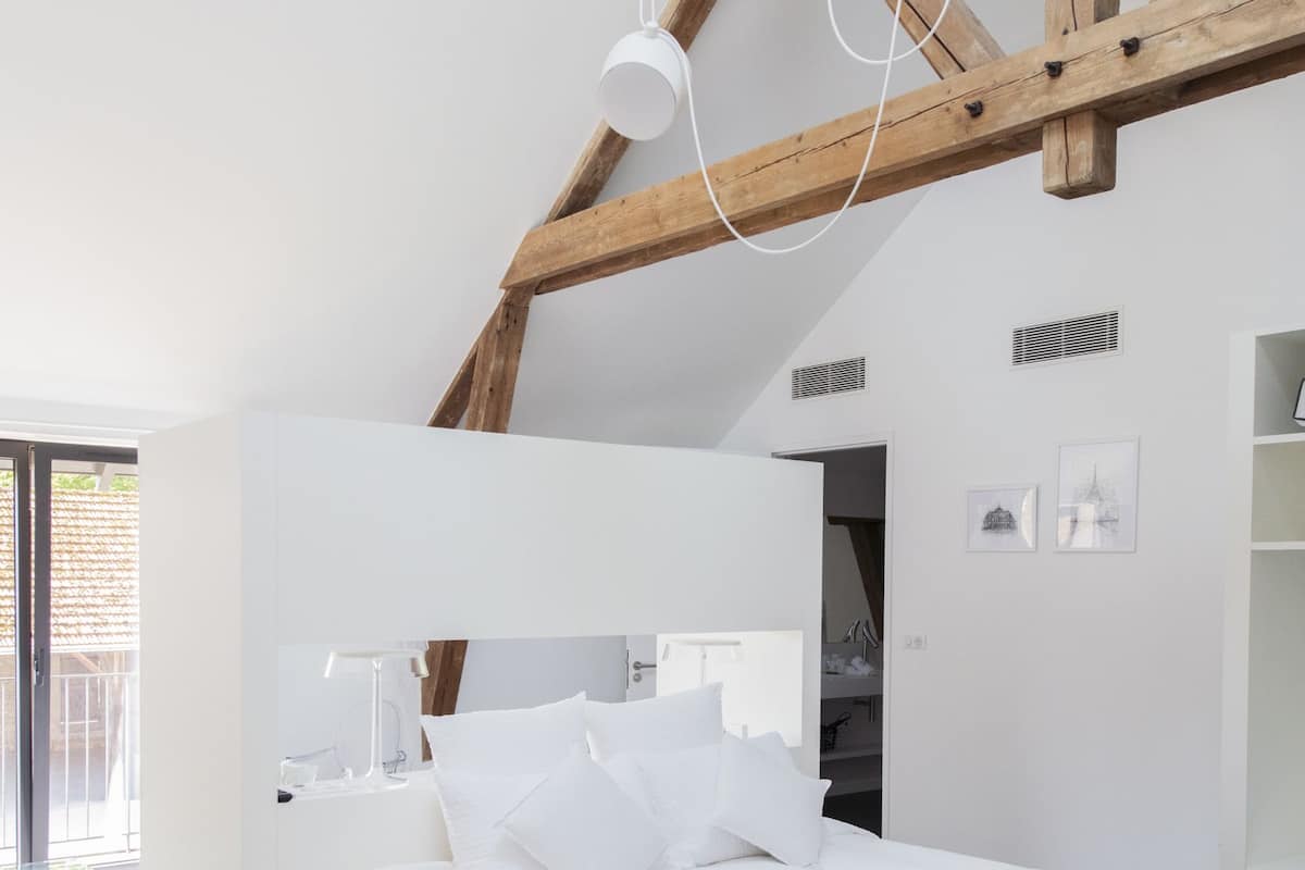 Quarto moderno com cama branca, vigas de madeira e uma janela grande.