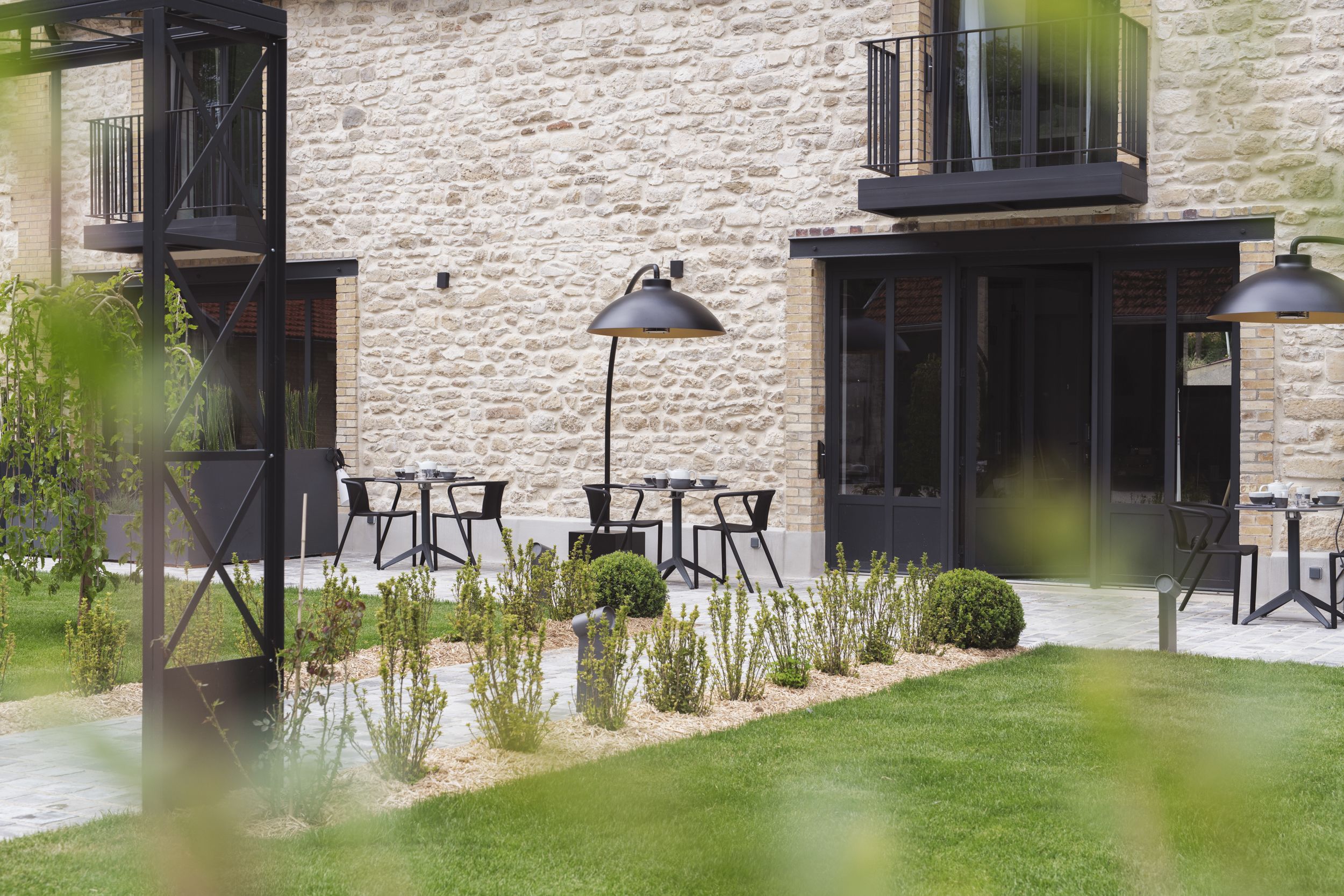 An outdoor seating area with tables and chairs, a stone building with a balcony, and a small garden with plants.
