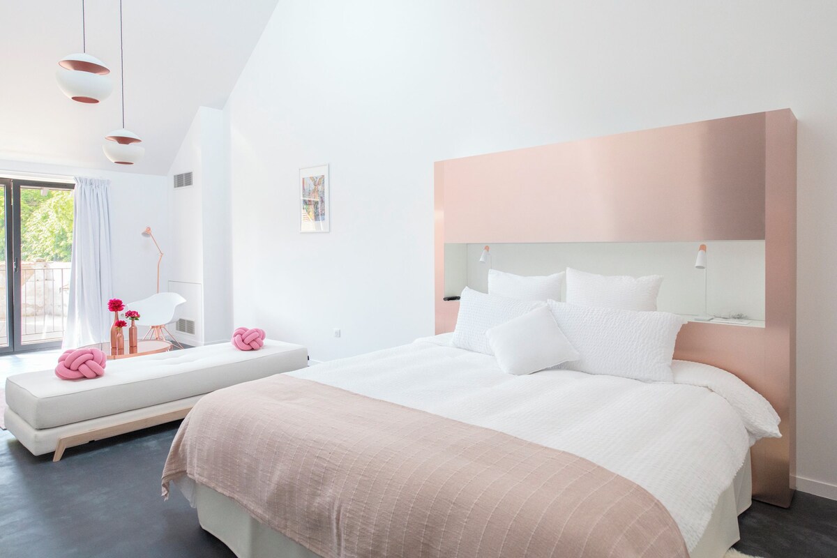 Un dormitorio moderno con una cama grande, una mesita de noche, un banco con objetos decorativos y una ventana con cortinas.