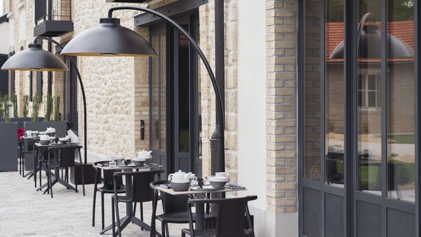 Un bâtiment doté d’une façade en pierre, de fenêtres à encadrement noir et d’un espace extérieur aménagé avec des tables et des chaises.