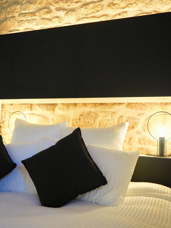 A bed with white and black pillows, a stone wall behind, and a nightstand with a lamp.