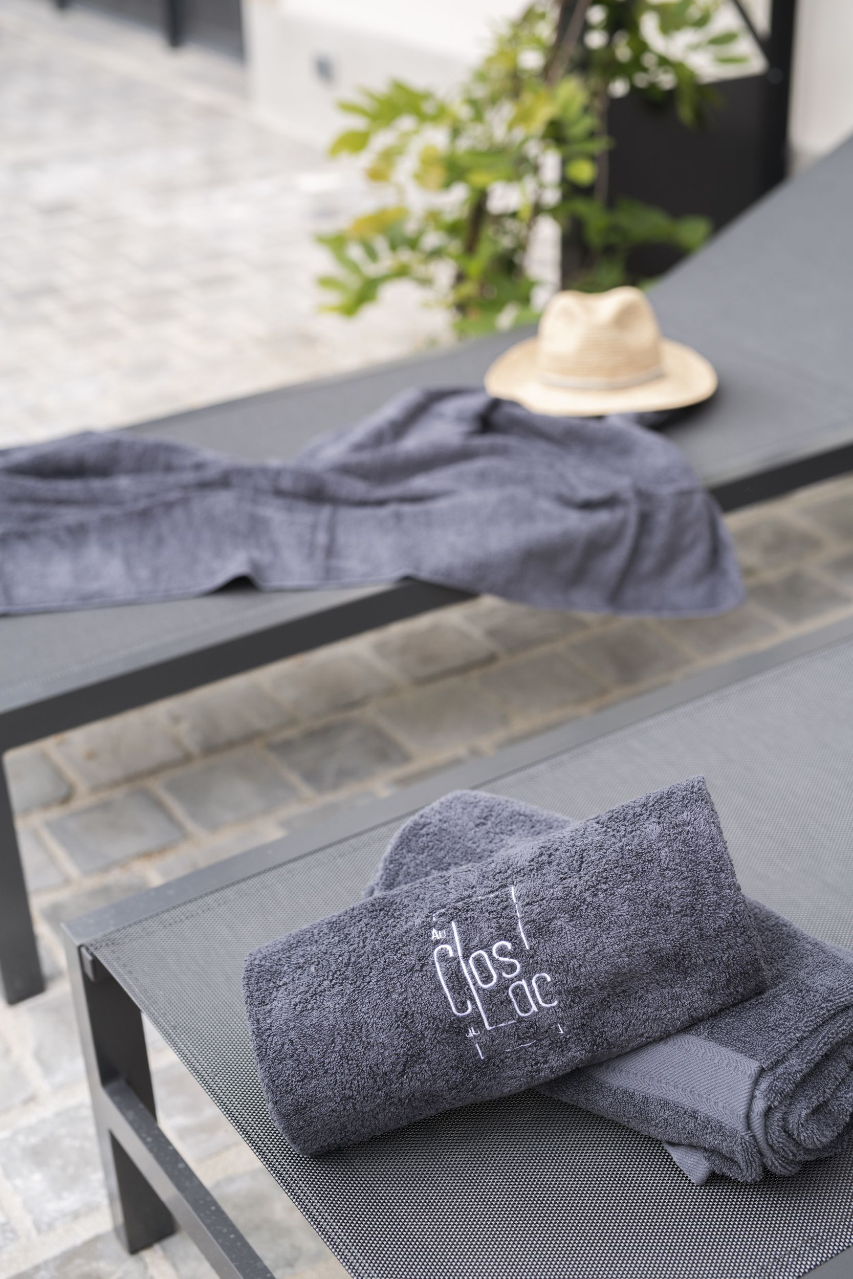 A neatly folded gray towel with "Clostac" embroidered on it, placed on a textured surface, likely a lounge area with a hat and a plant in the background.