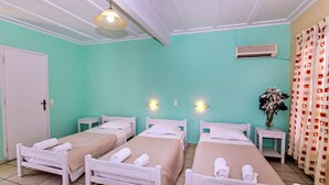 Habitación con tres camas, cada una con cabecera blanca y colcha color beige. Hay dos mesitas de noche, una puerta blanca y una pequeña mesita auxiliar blanca con un jarrón de flores.