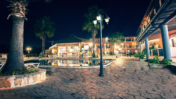 Una escena nocturna de un patio con piscina, palmeras y un edificio de varios pisos y balcones.