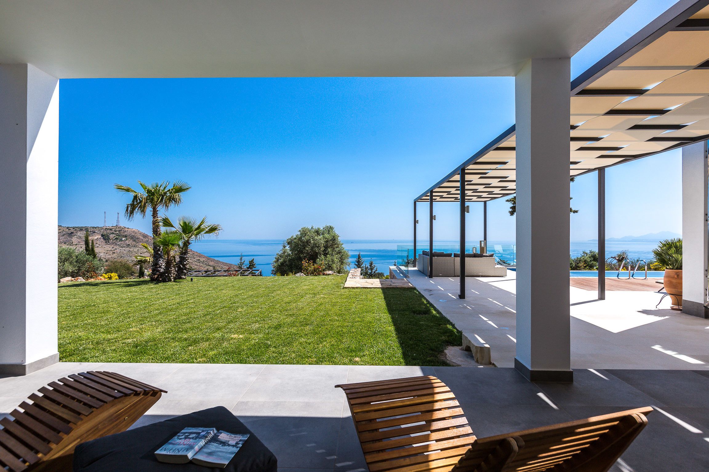 A modern outdoor patio with a view of the ocean, featuring a grassy lawn, wooden benches, and a pergola structure.